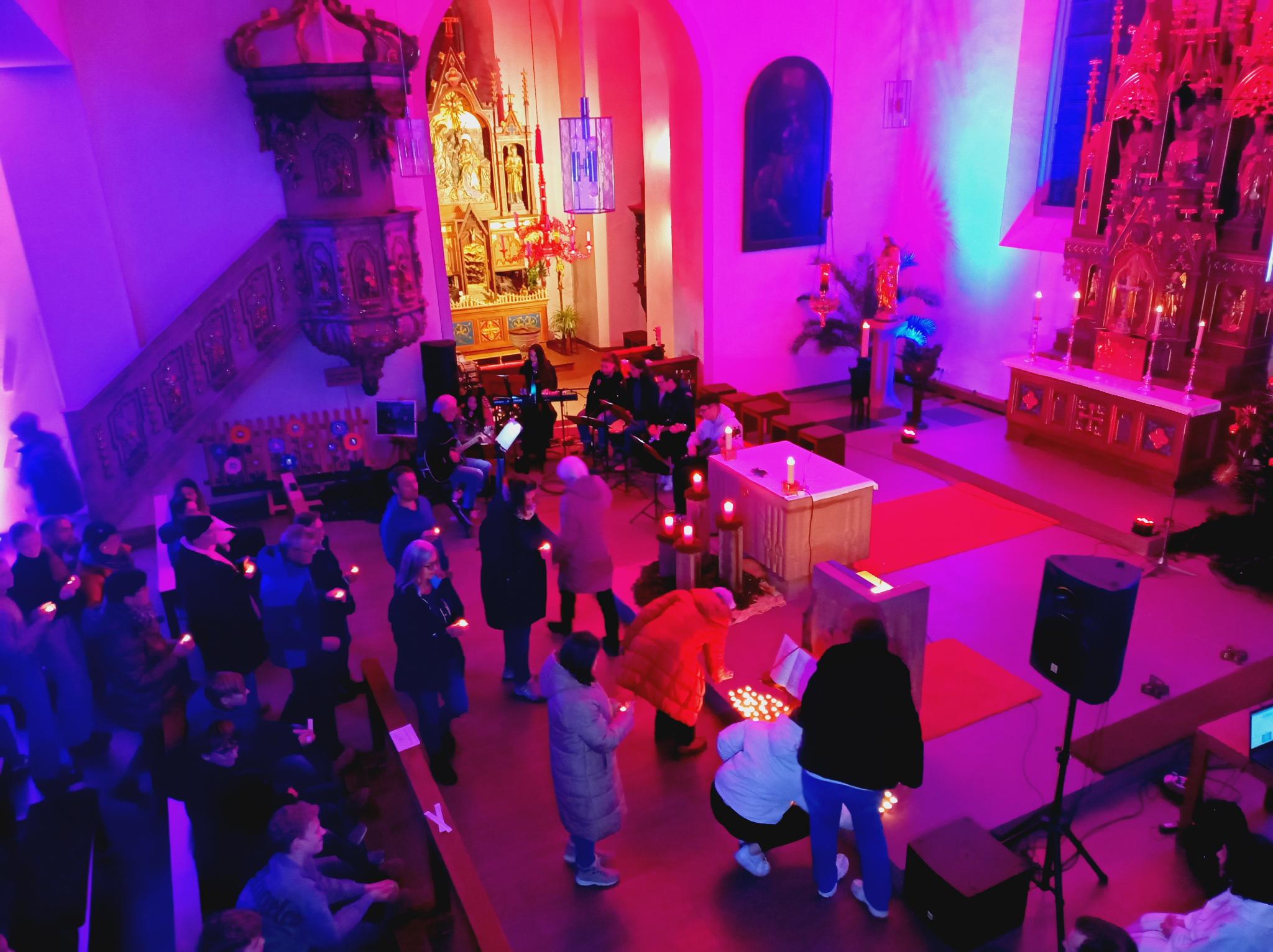 Blick von der Empore in den Kirchenraum. Die Besucher halten ein brennendes Teelicht in der Hand und bringen es nach vorne, um es beim Evangelium abzustellen. Dabei spielt die Band im Hintergrund Musik.