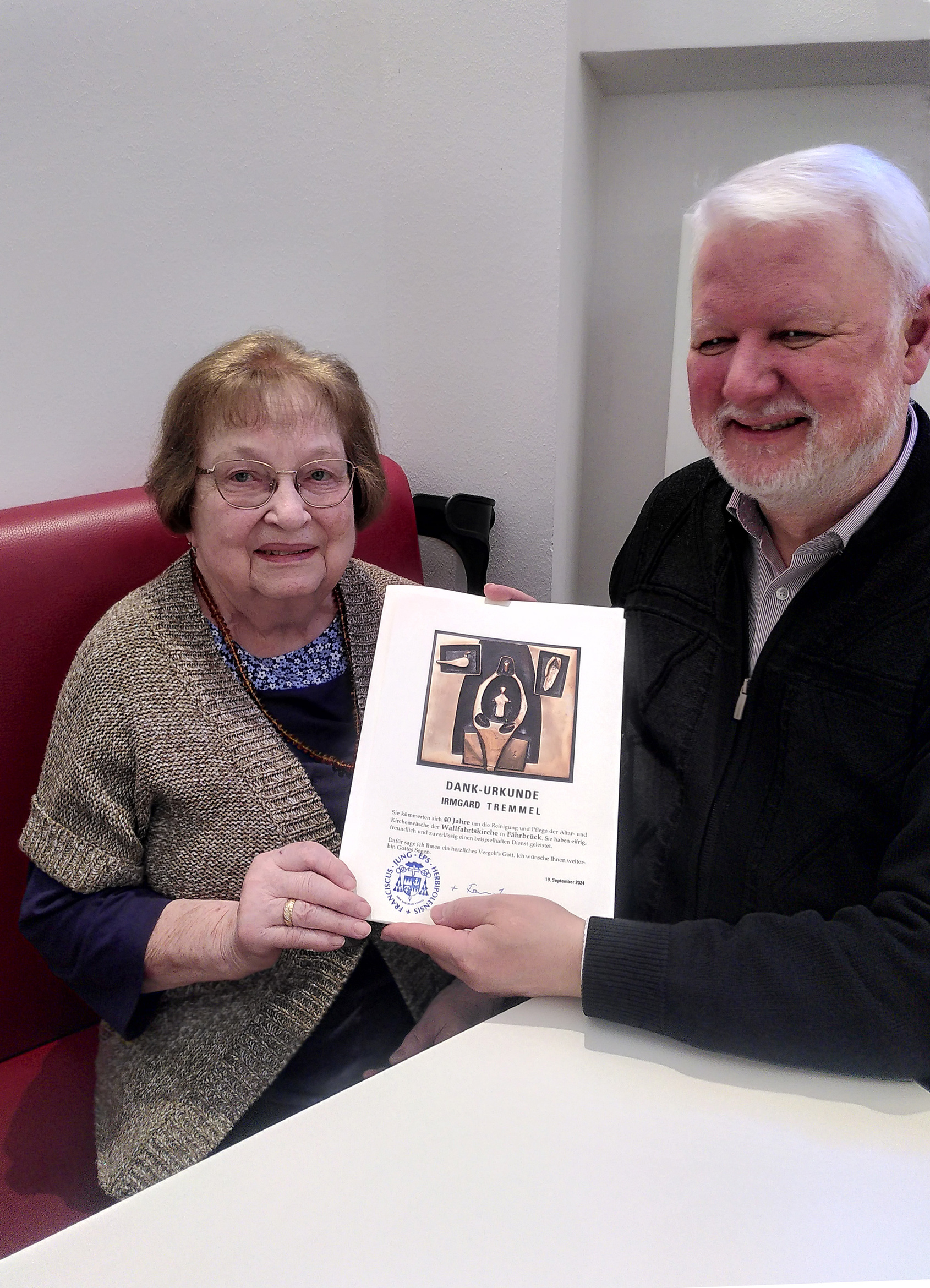 Frau Tremmel und Pater Matthäus sitzen an einem Tisch und zeigen in der Mitte die Dankurkunde des Bischofs.