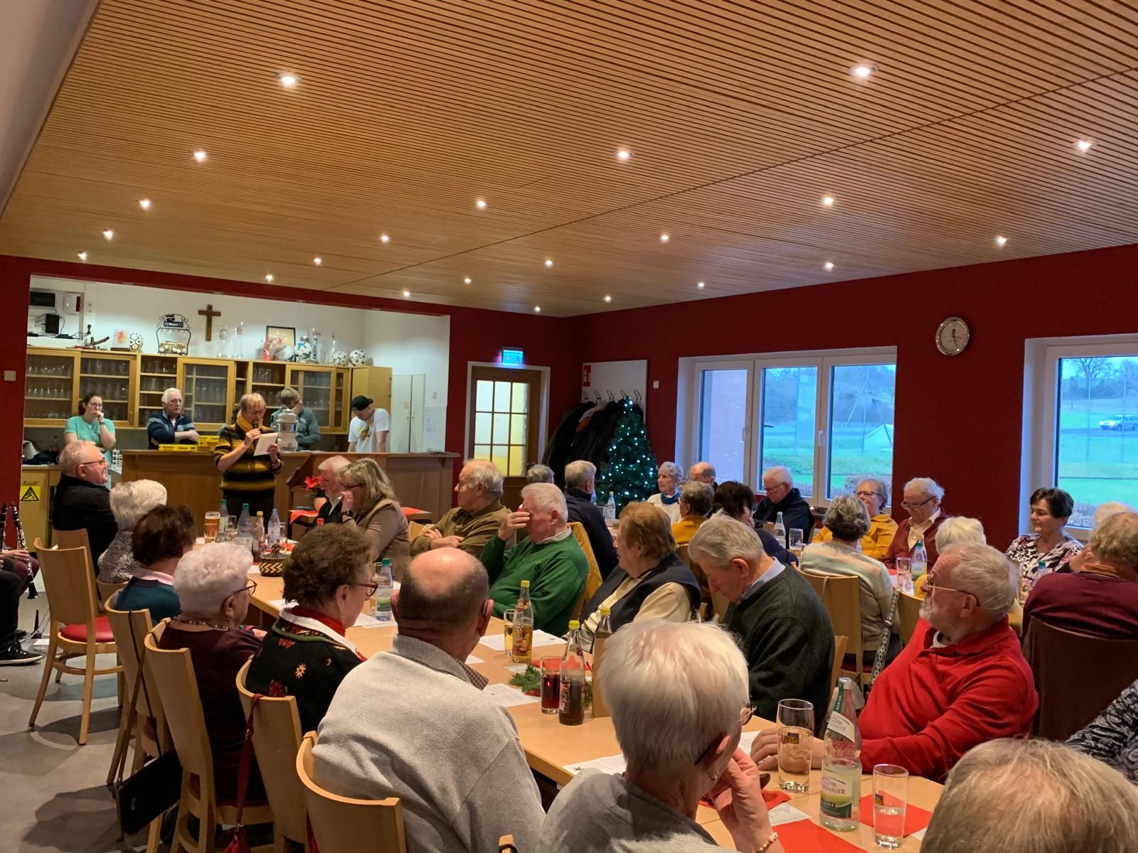 Blick von hinten auf den gut gefüllten Saal des Sportheimes, vorne steht eine Frau und trägt einen Text vor