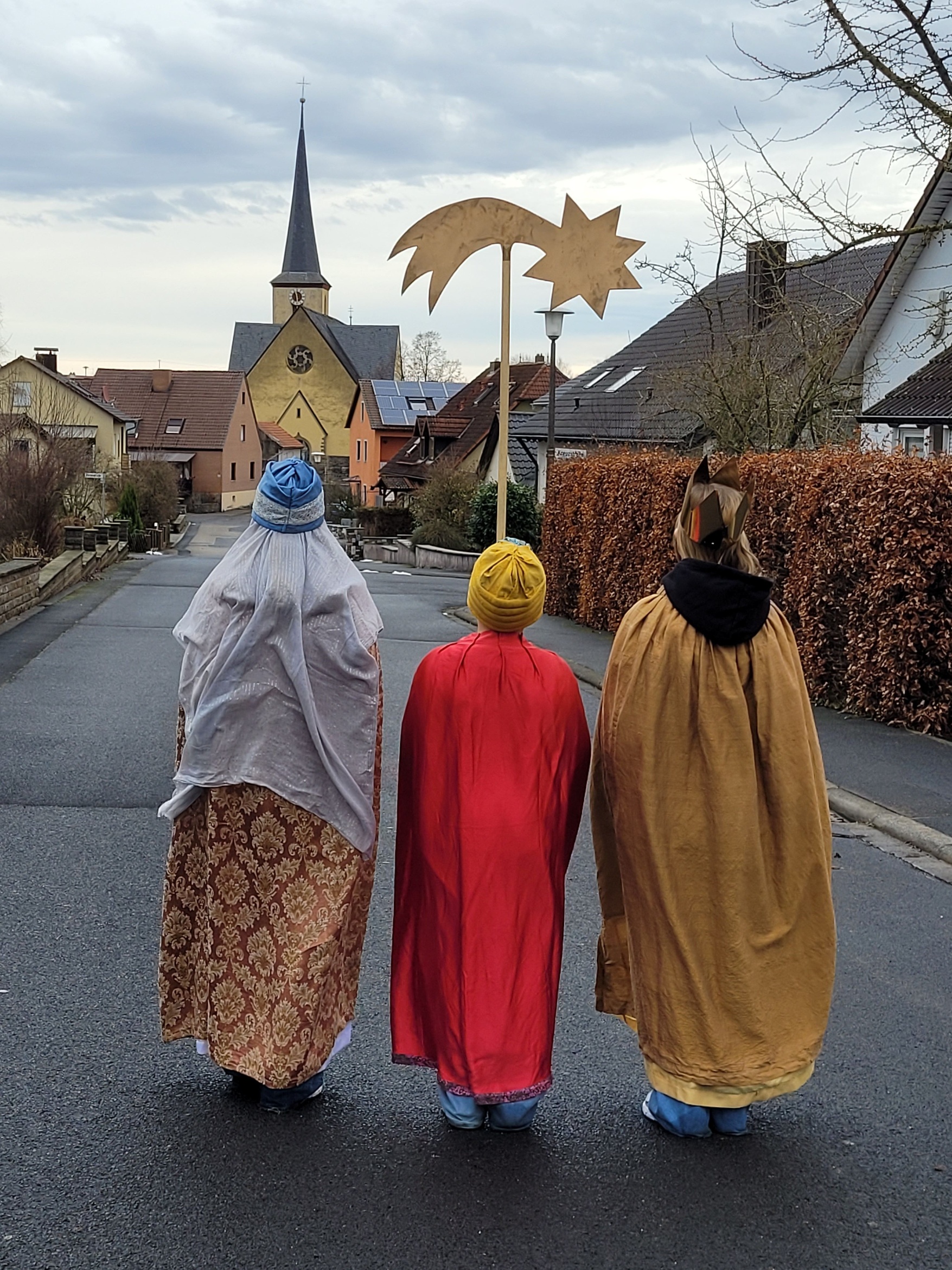 Blick von hinten auf drei Sternsinger, der mittlere hält einen Stern hoch. Im Hintergrund sieht man die Kirche.