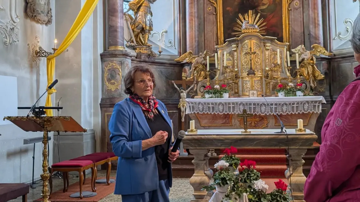 Eine Frau steht mit einem Mikrofon in der Hand neben dem Altar einer barocken Kirche.
