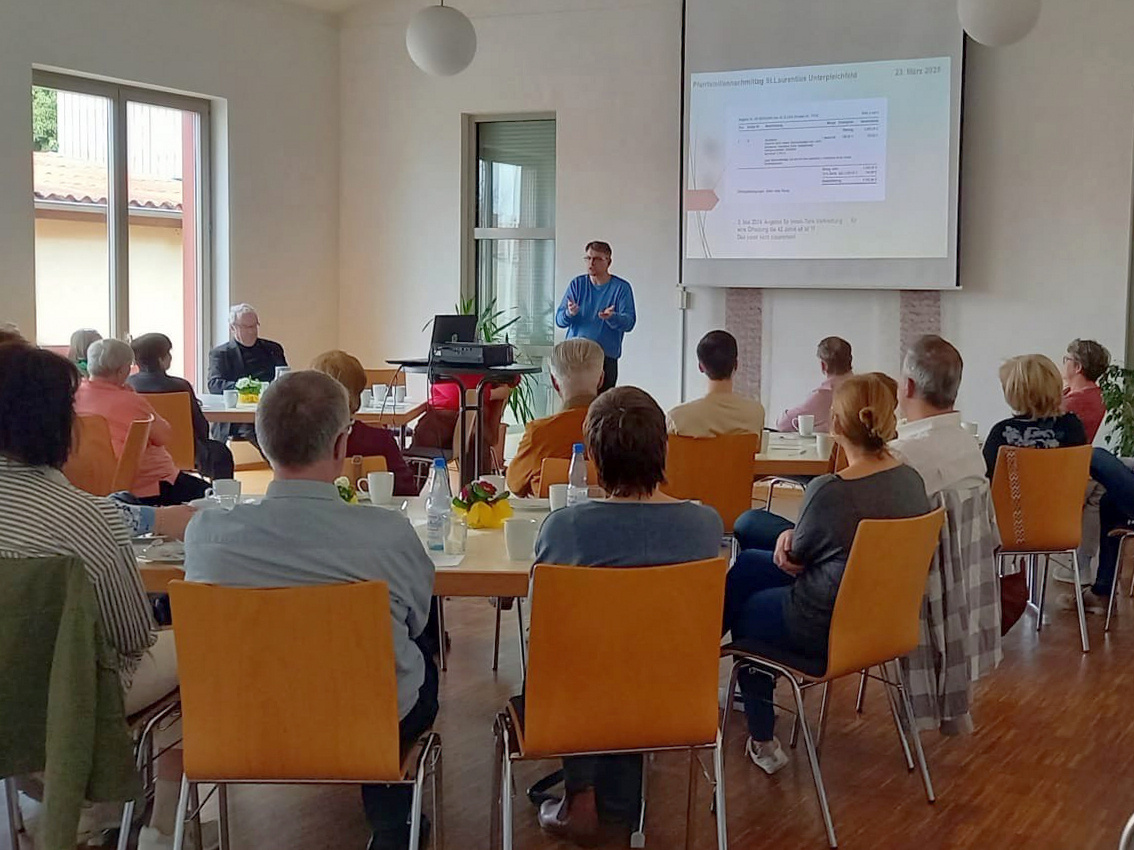 Ein schönes helles Bild mit Blick in das Pfarrheim. Auf Stühlen sitzen die Menschen und schauen nach vorn zum Redner.