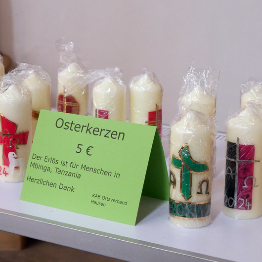Verschiedene Osterkerzen stehen auf einem Tisch. Davor ist ein grünes Schild. Darauf steht, wofür der Erlös der Aktion gedacht ist.