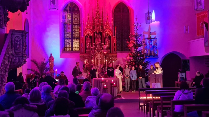 Blick auf den Altarraum der Kirche. Die Kirche ist mit Strahlern lila und rosa erleuchtet, im Altarraum stehen einige Jugendliche und lesen etwas vor.