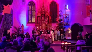 Blick auf den Altarraum der Kirche. Die Kirche ist mit Strahlern lila und rosa erleuchtet, im Altarraum stehen einige Jugendliche und lesen etwas vor.