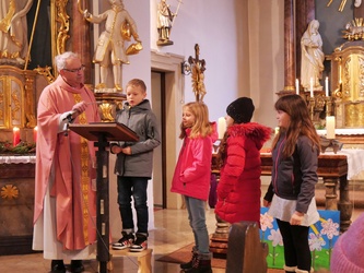 Pfarrer Rügamer steht links. Rechts neben im sind vier Kinder. Ein Junge spricht gerade ins Mikrofon.