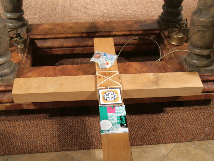 Ein Holzkreuz liegt auf dem Boden. In der Mitte sind Gegenstände angeheftet.