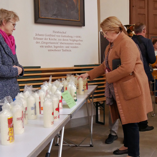 Auf einem Tisch stehen mehrere Osterkerzen. Links vom Tisch ist Frau Altenhöfer. Rechts vom Tisch steht eine Frau und schaut sich die Kerzen an.