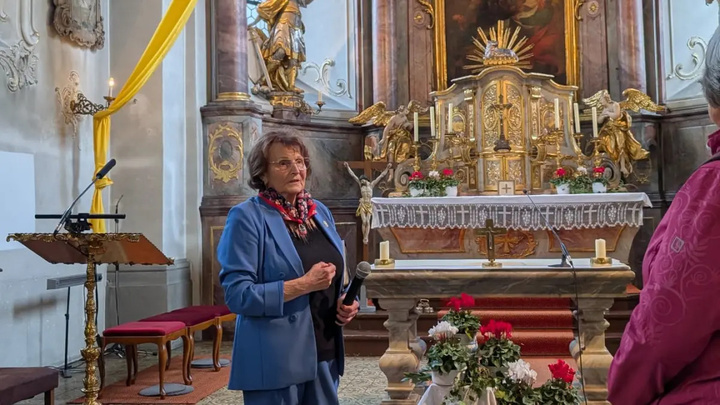 Eine Frau steht mit einem Mikrofon in der Hand neben dem Altar einer barocken Kirche.